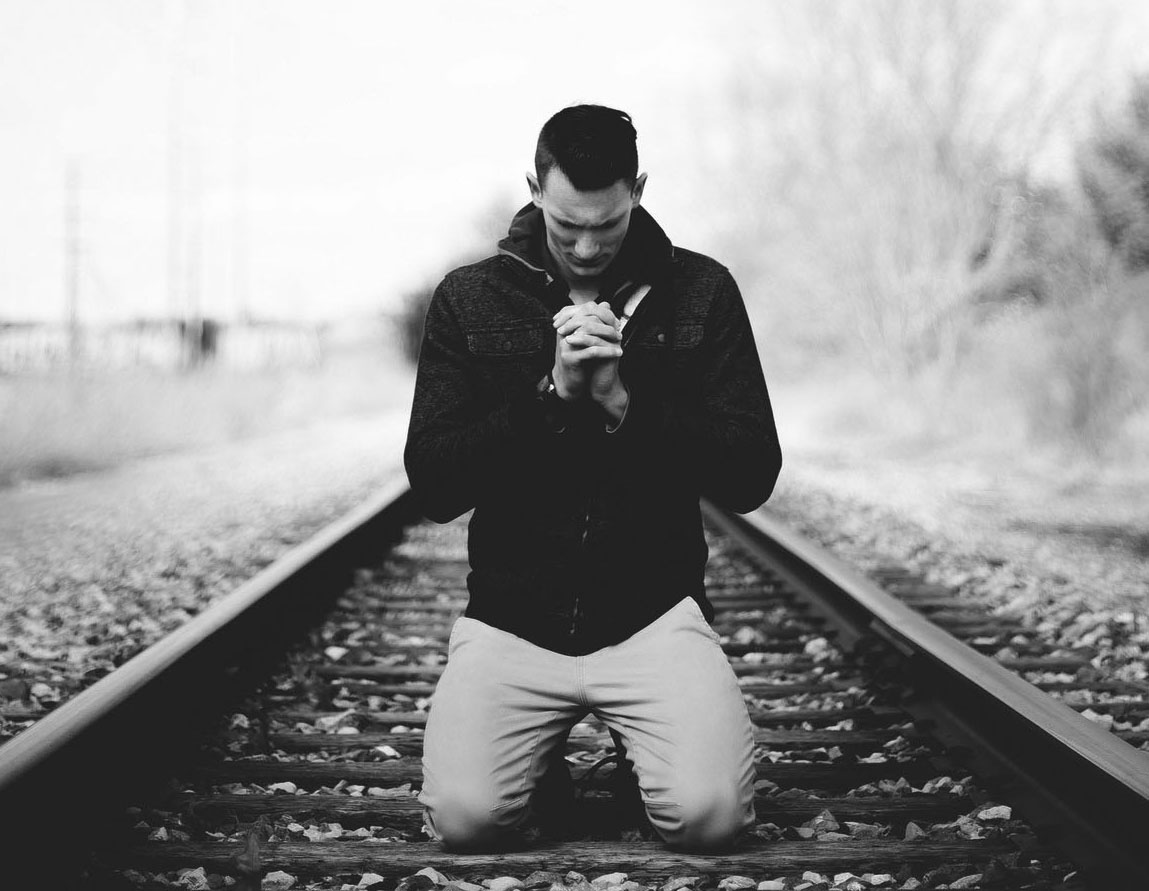 A corcasian man dressed in a black jacket and grey pants kneeling on a train tracks in the prayer position.